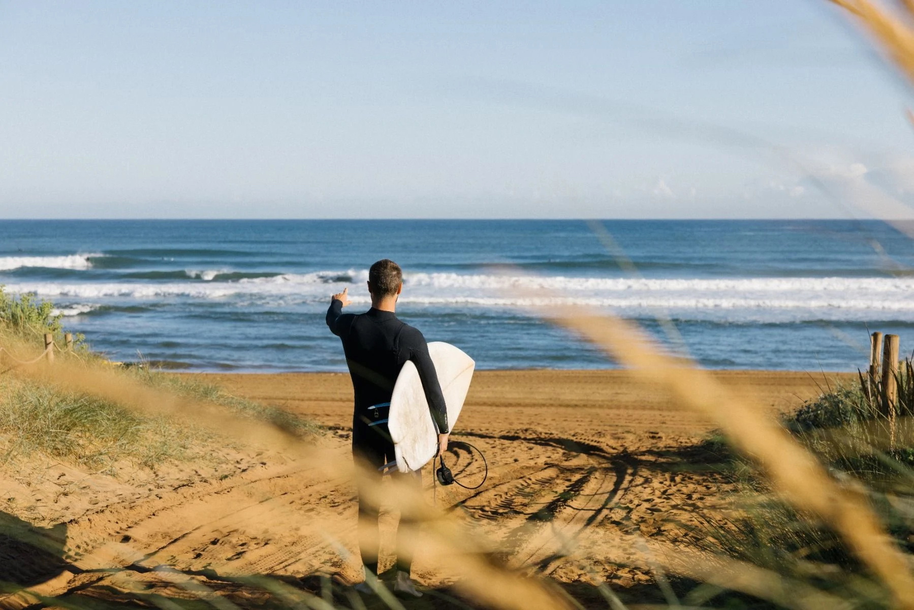 Surfer in een zwart wetsuit loopt met de plank onder zijn arm over een zandig duinpad naar het strand