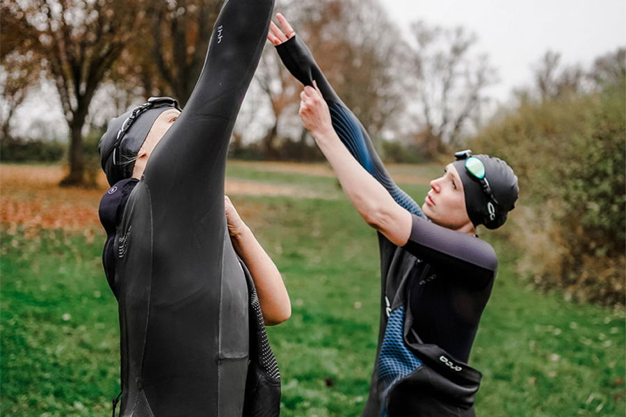 Twee zwemmers in wetsuit en badmuts op het gras, de een helpt de ander na het zwemmen uit het pak