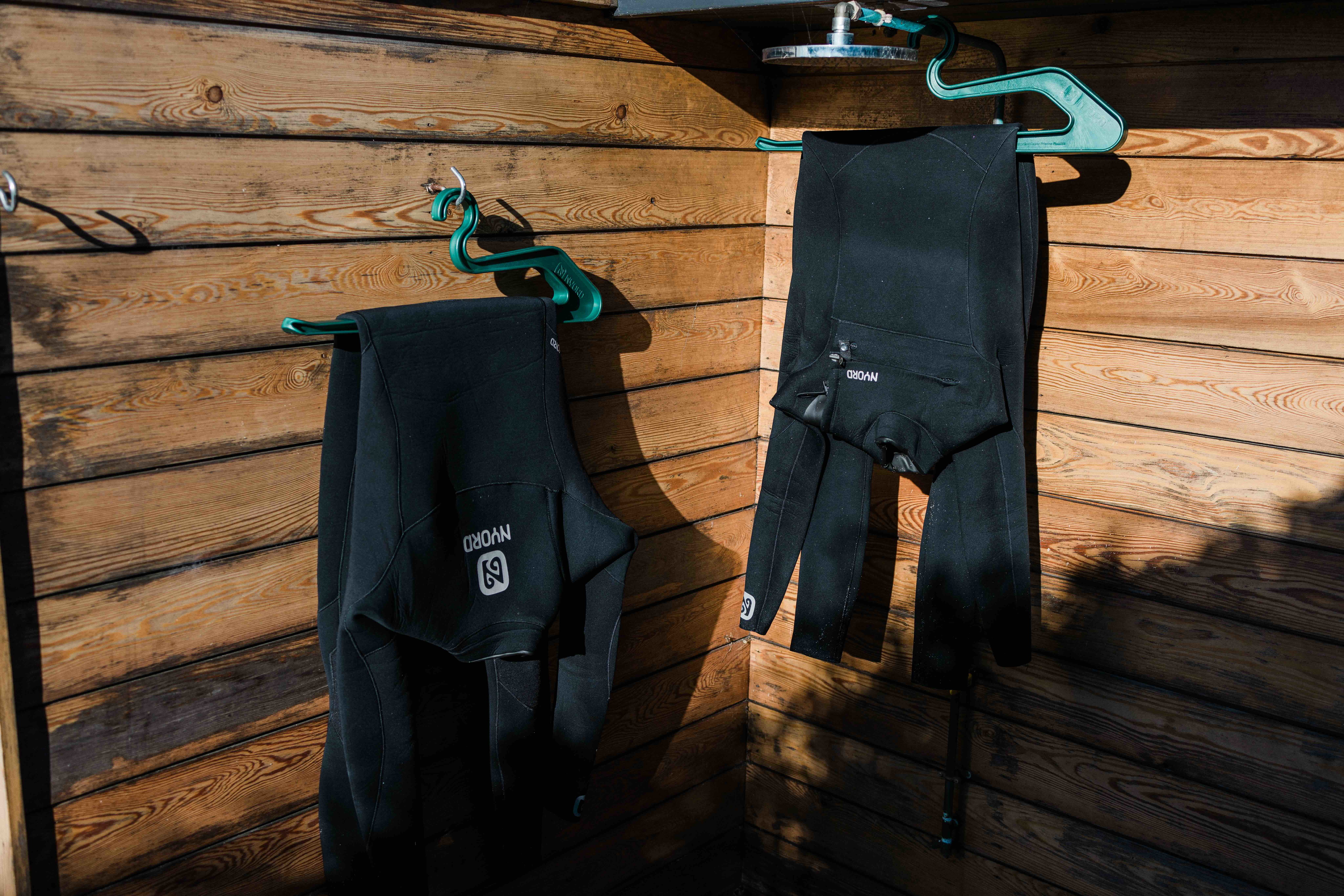 Twee zwarte wetsuits hangen te drogen aan hangers onder een houten strandhuisje