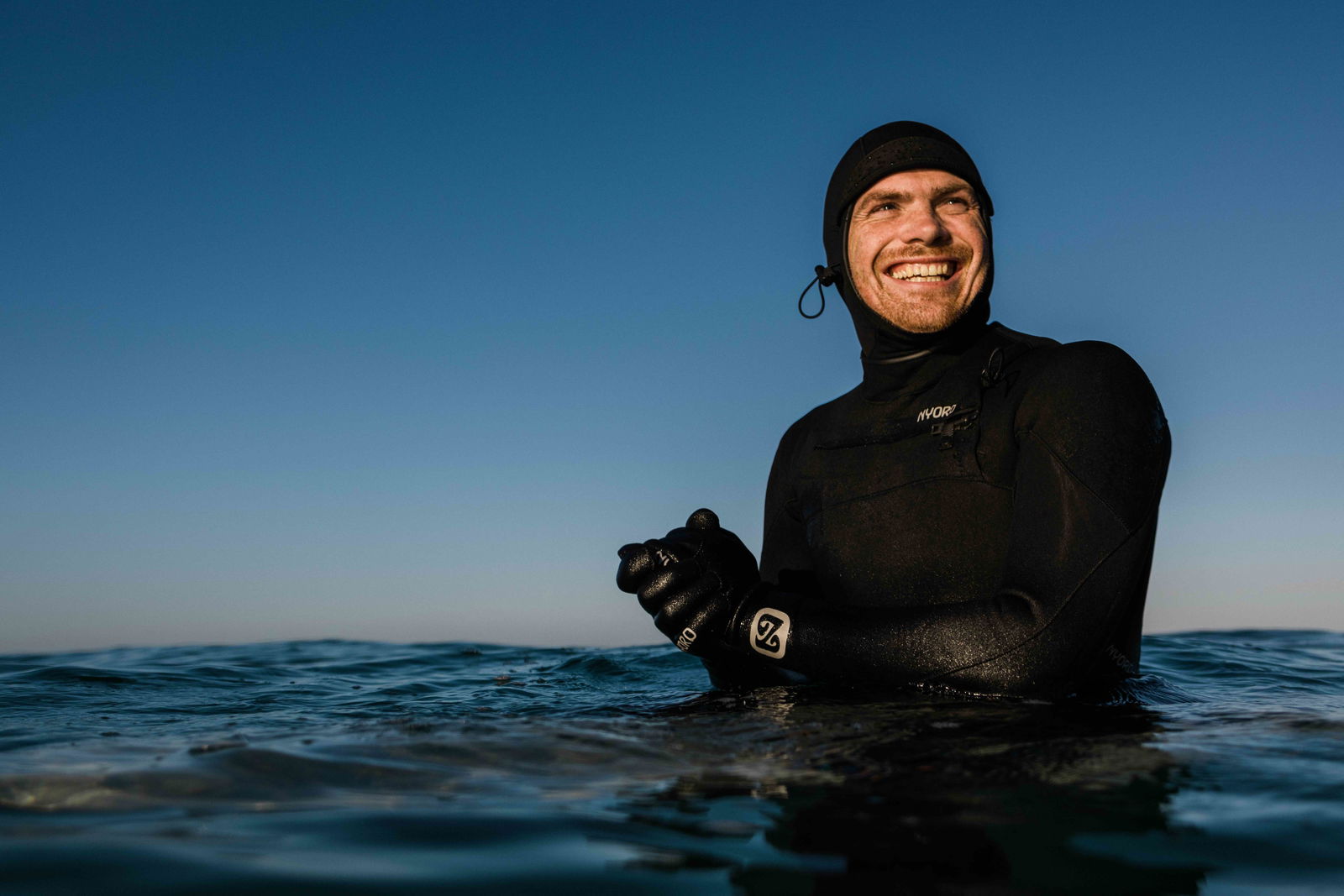 Lachende man in een zwart wetsuit met hoofdkap en handschoenen tot borsthoogte in zee
