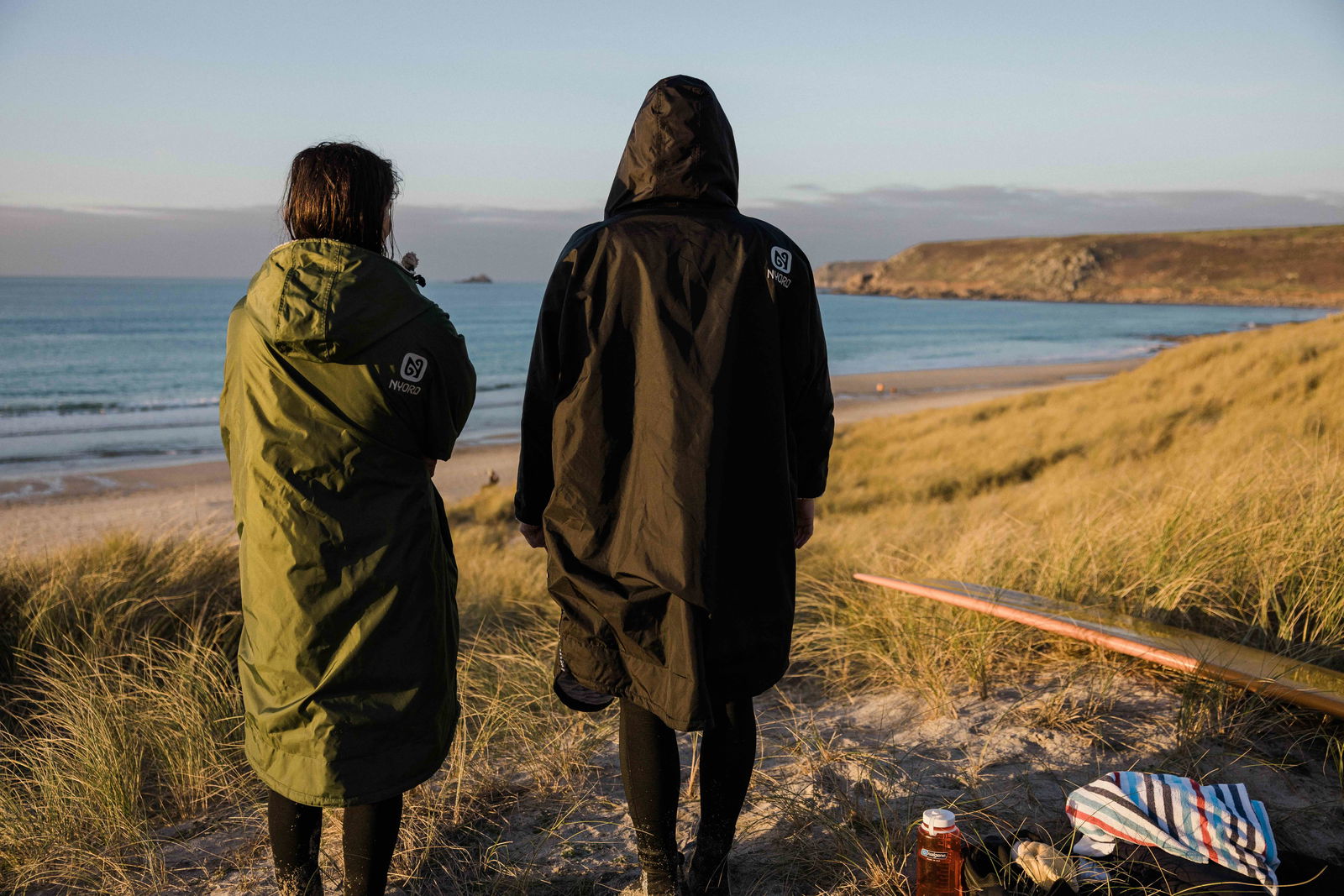 Twee mensen in lange omkleedjassen met capuchon kijken bij zonsondergang uit over het strand