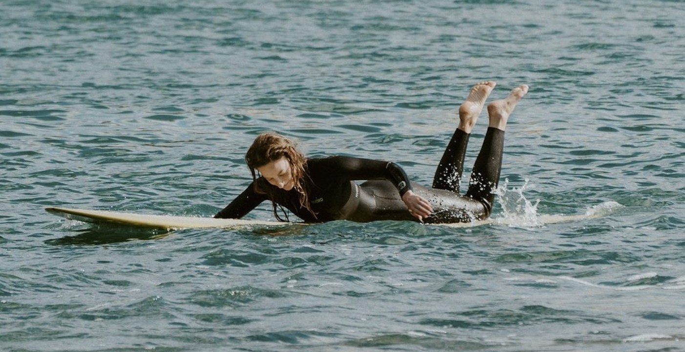Twee surfers in zwarte wetsuits peddelen samen op een longboard de zee in
