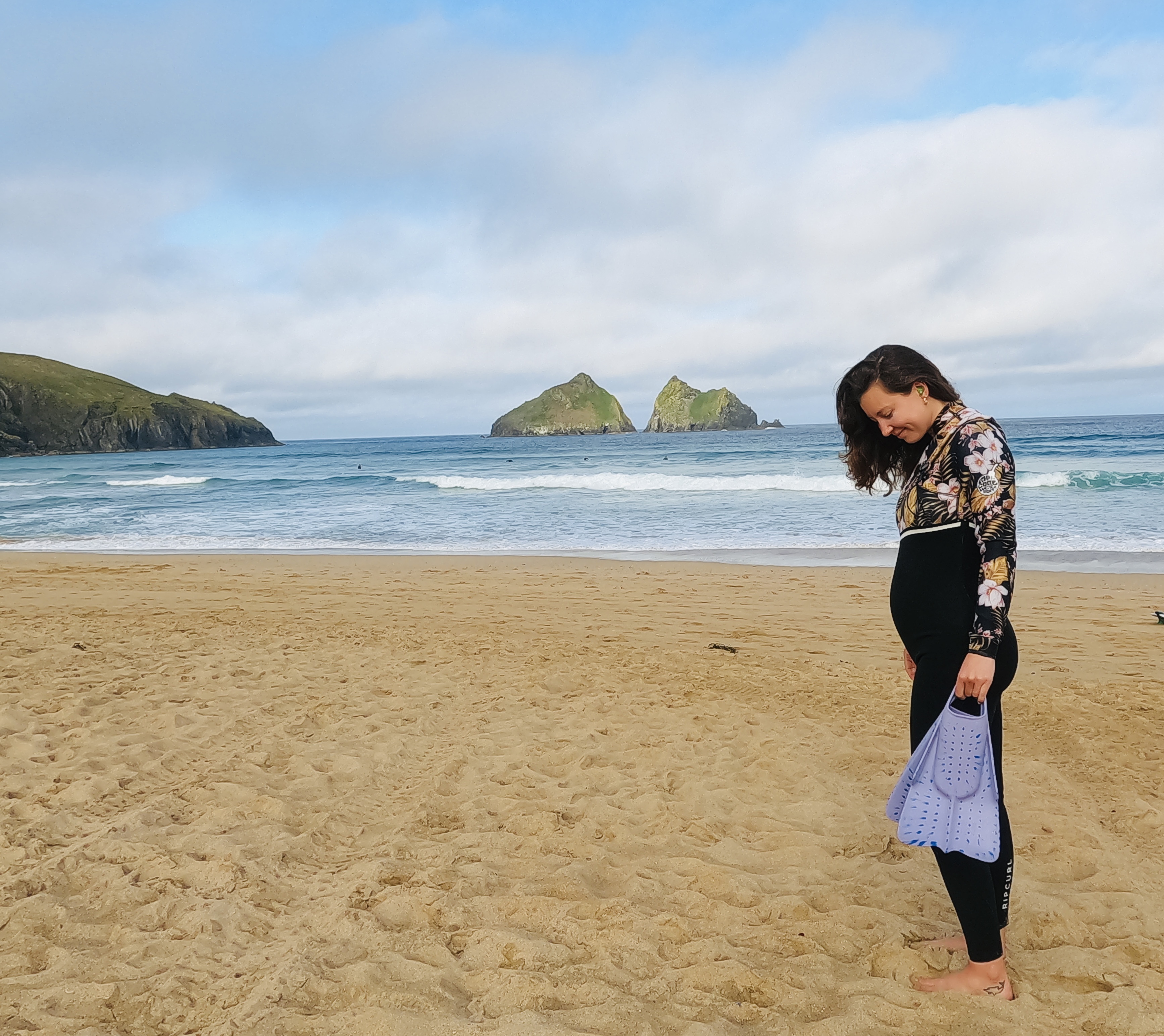 Pregnant surfer on a sandy beach in a patterned long-sleeved top and leggings, holding swim fins