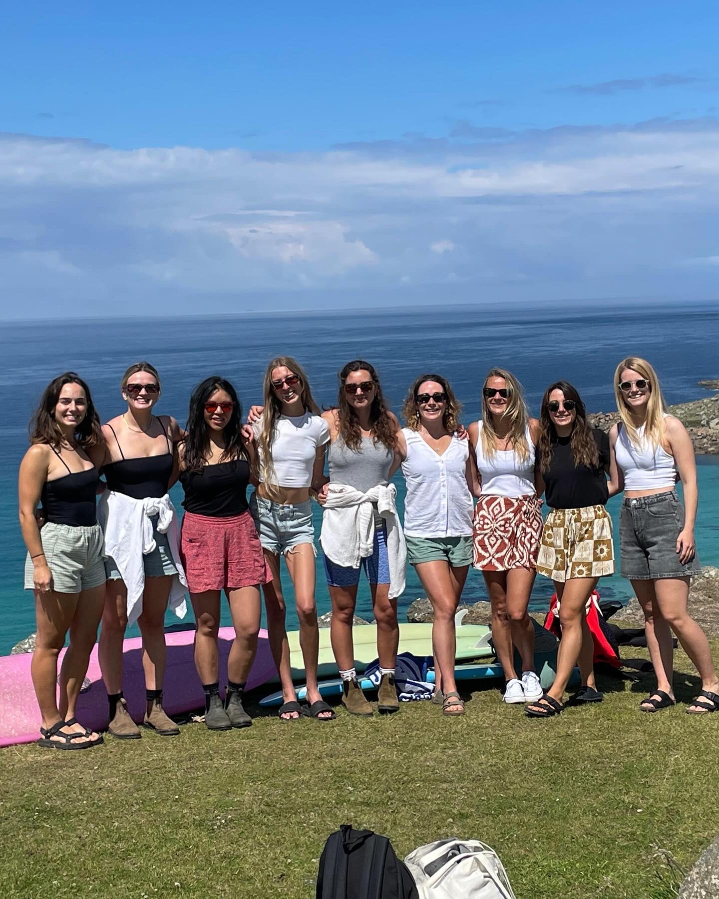 Group of women standing together on the grass above a beach with surfboards laid out behind them