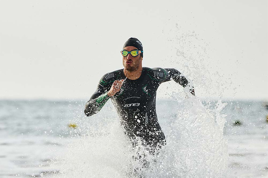 Man in een ZONE3 zwemwetsuit die door de branding waadt, omringd door opspattend water
